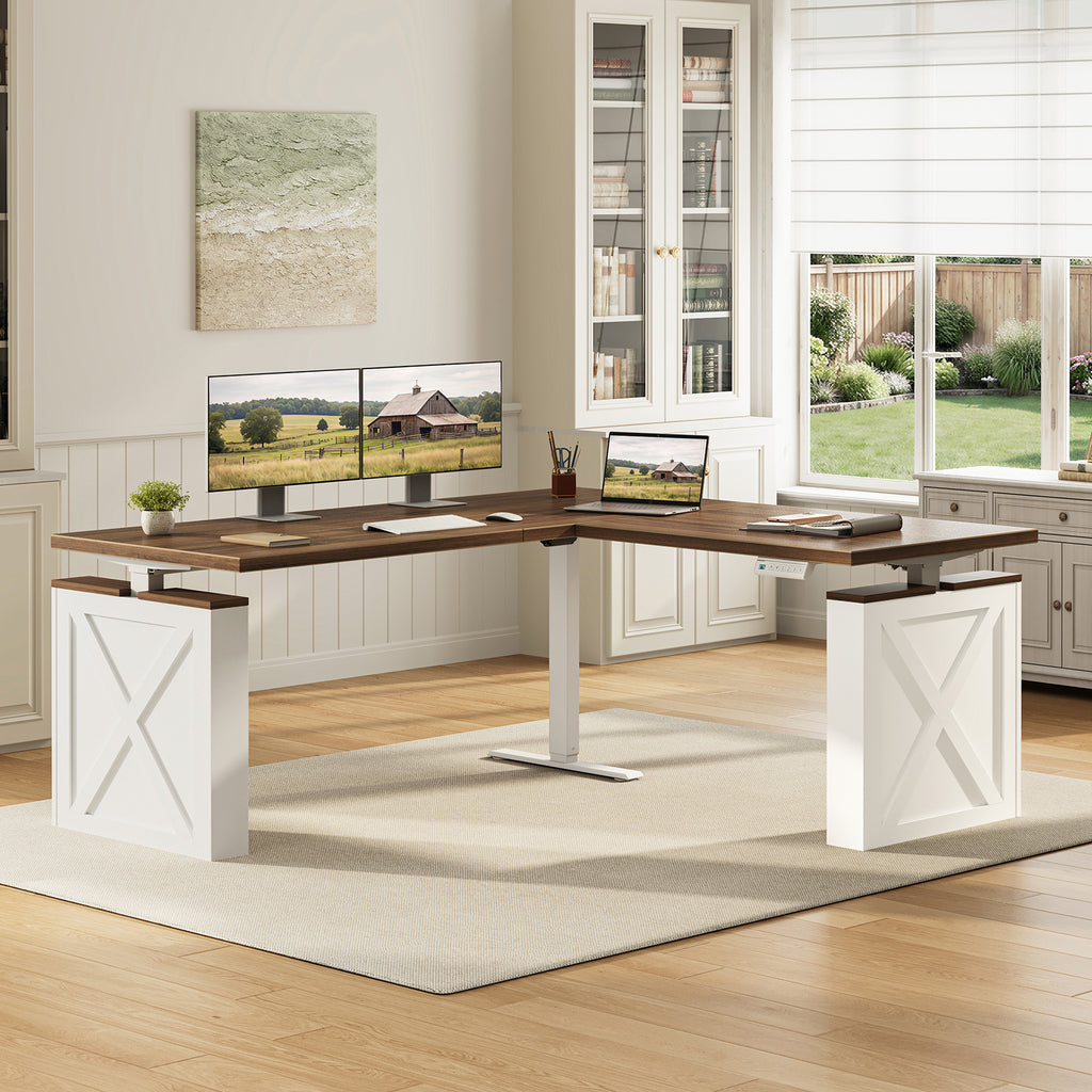 OffiGo 63-inch farmhouse L-shaped standing desk with a walnut-brown desktop, white X-brace side panels and a white center support leg, arranged in a bright home office with dual monitors, a laptop and a printer beside white bookcases.
