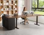 Home office scene showing the OffiGo 71-inch executive L-shaped standing desk with a walnut-brown desktop, black legs and a rolling black storage cabinet, with a seated user at dual monitors beside a bright window.