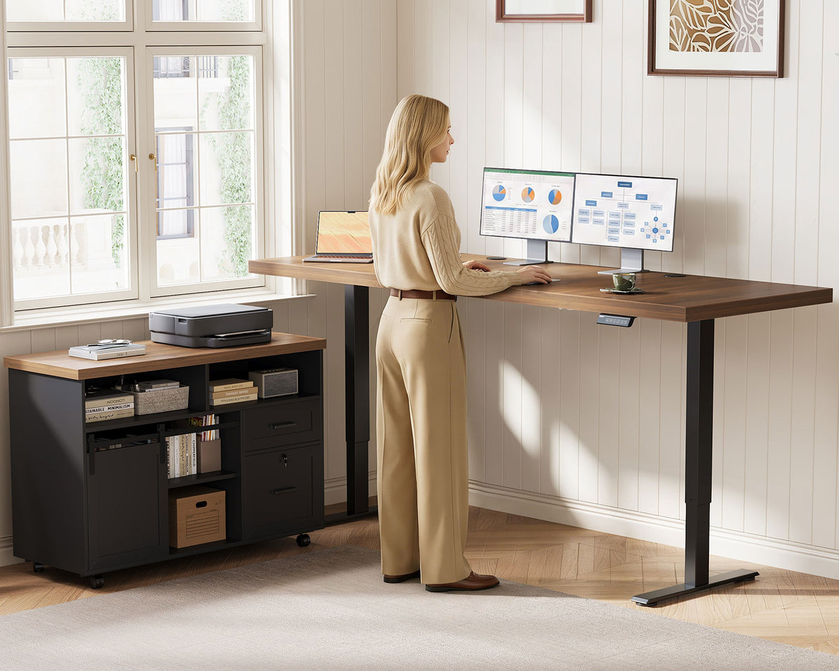 Standing user working at the OffiGo 71-inch executive L-shaped standing desk with a walnut-brown desktop, black legs and a rolling black storage cabinet, with dual monitors and a printer in a sunlit home office.