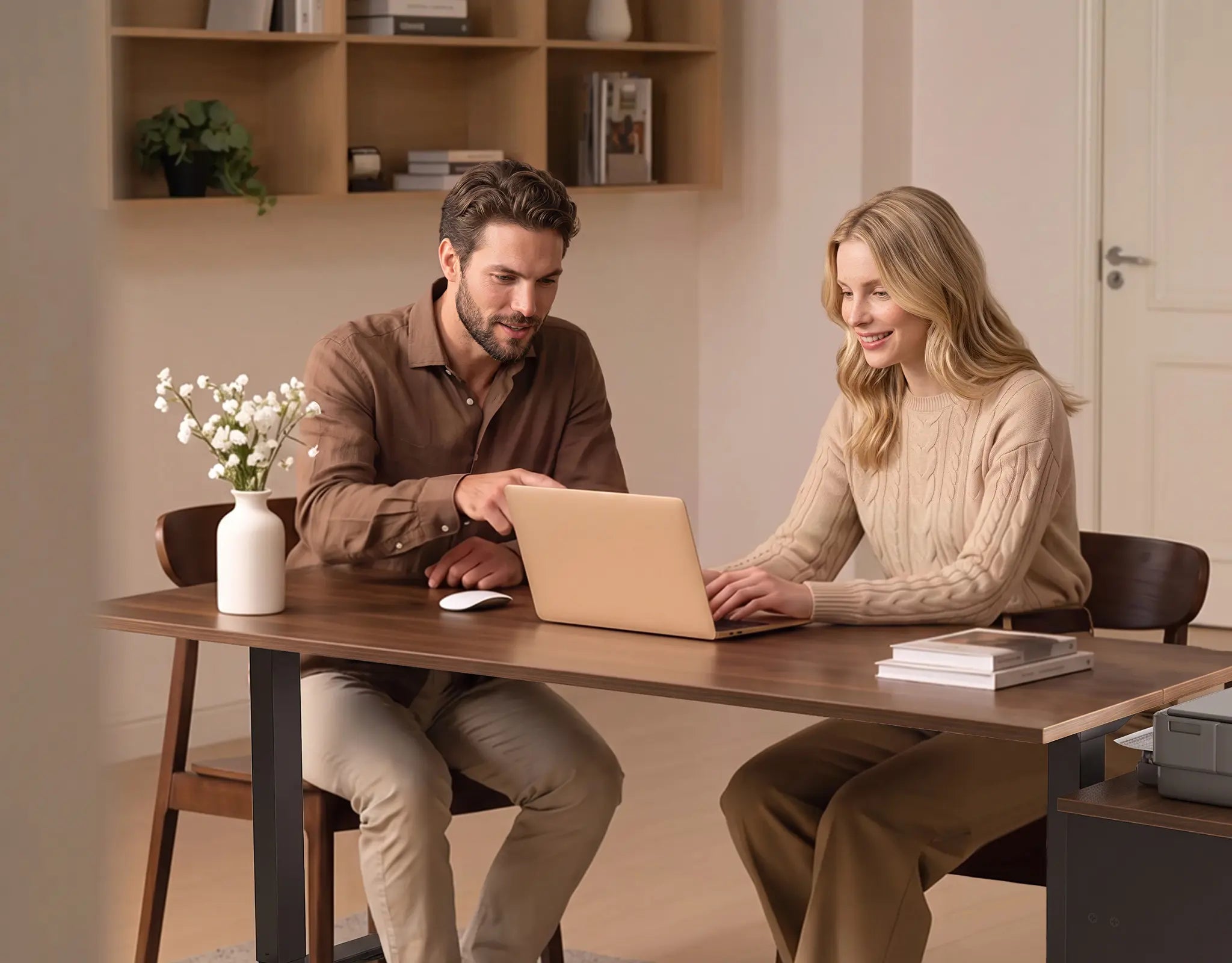 Two people seated together at an OffiGo wood-look desk, using a laptop beside a vase of flowers and books.