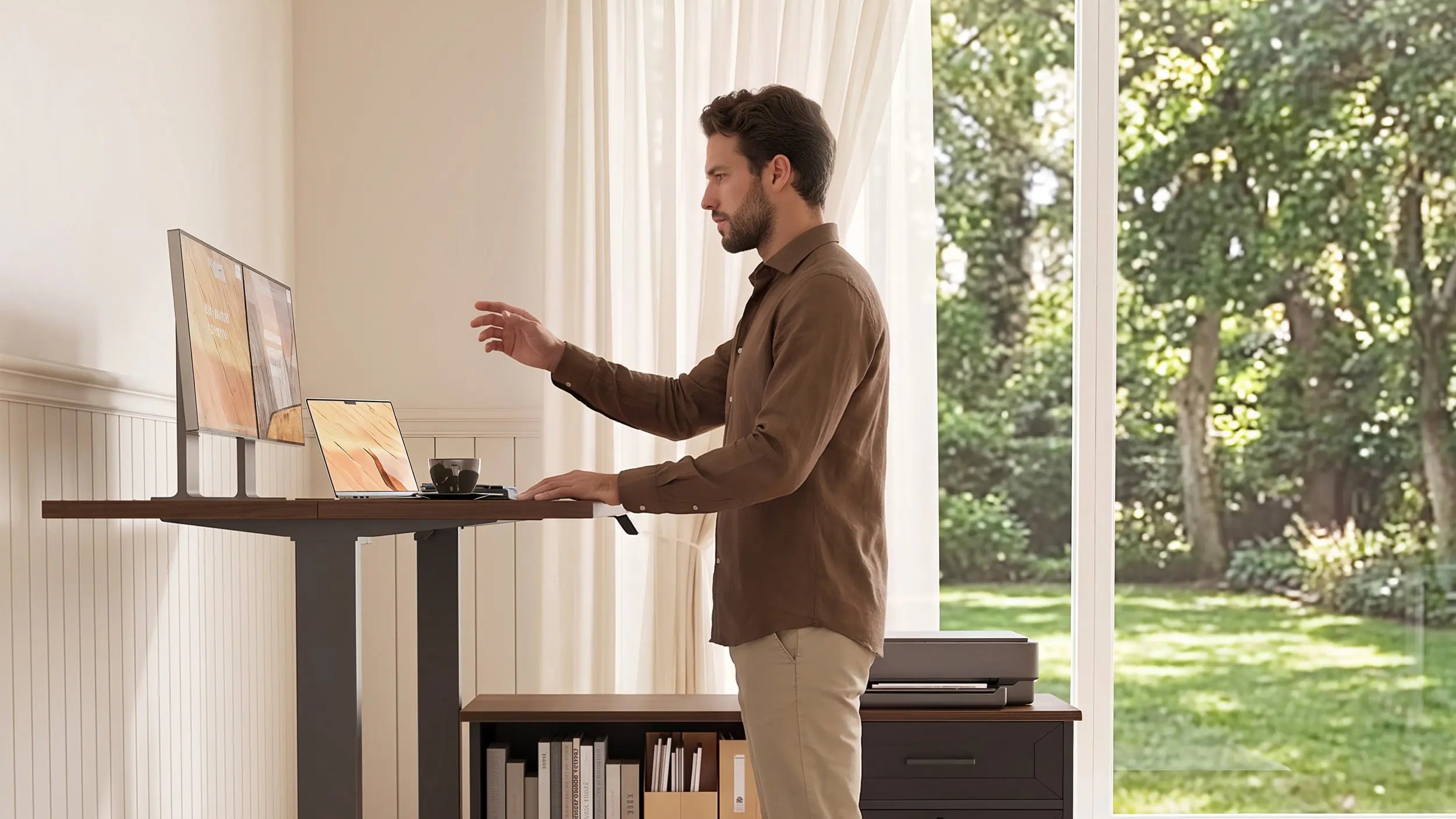 A man working at a raised OffiGo standing desk with a laptop and monitors, beside a rolling storage cabinet and a garden-facing window.